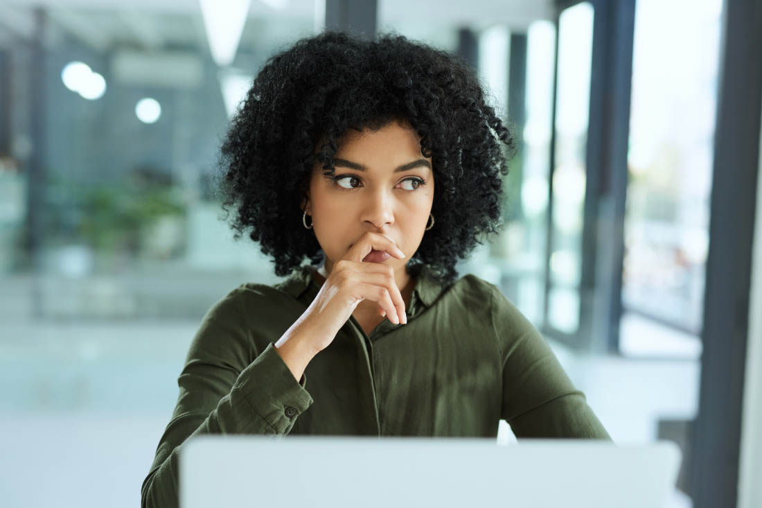Woman sitting at a desk looking thoughtful, with one hand resting on her chin and a laptop in front of her.