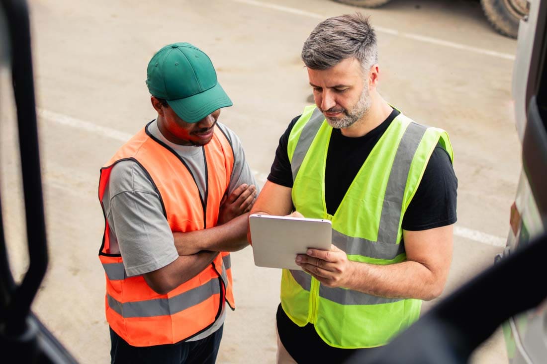 Two workers wearing safety vests discussing logistics while reviewing a tablet outdoors at a transportation site.