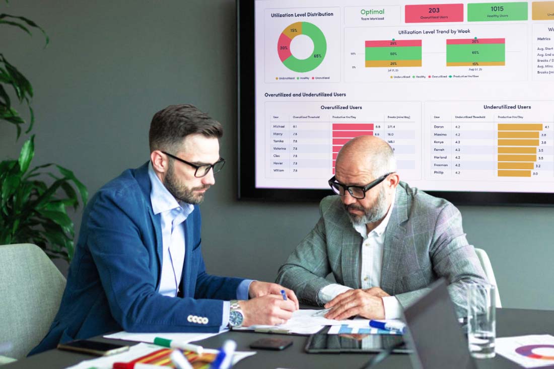 Two people reviewing documents in a meeting room with a data dashboard in the background.