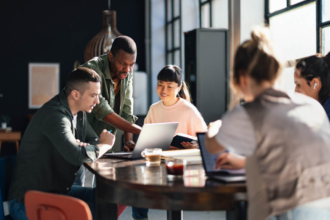 Diverse group of colleagues collaborating around a laptop in a modern, sunlit office space.