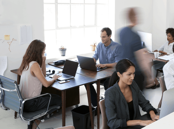 Several people working at desks in an office. A blurry man is walking by.